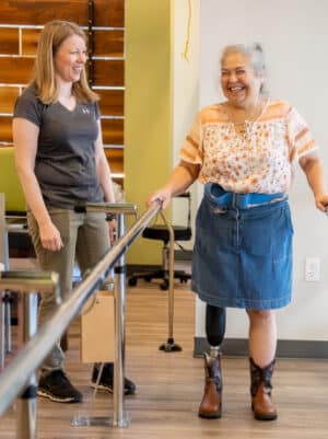 Woman with prosthetic leg smiling during physical therapy session with support from a caregiver