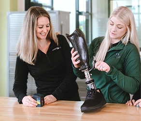 Two women adjusting a prosthetic leg with a shoe on a wooden table in a bright room