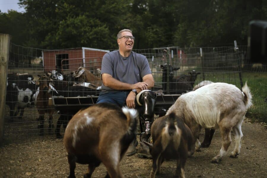Man laughing with goats on a farm with a red barn in the background