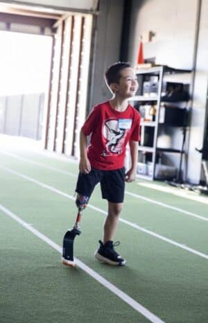 Young boy with prosthetic leg smiling on indoor track wearing red shirt and shorts