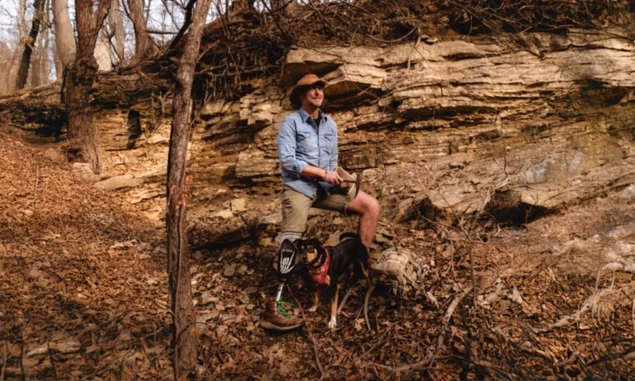 Man with a prosthetic leg hiking with dog in a forest rocky area in autumn.