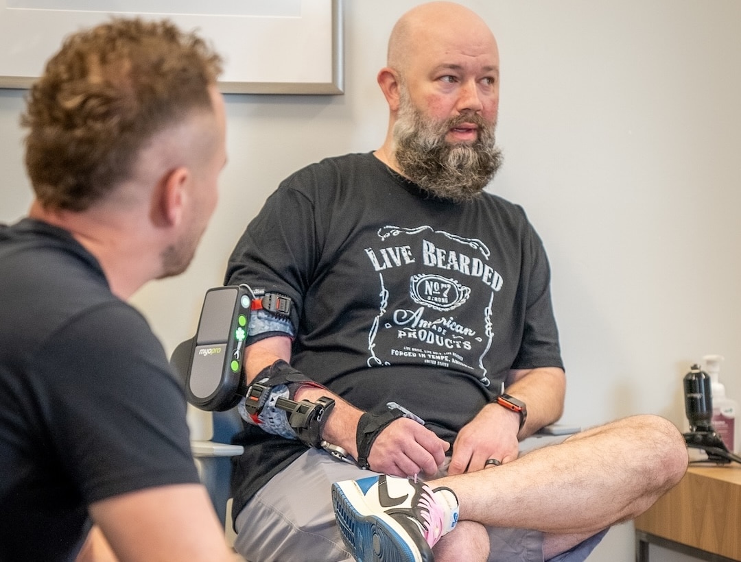 Man with a prosthetic arm sits indoors talking with another person